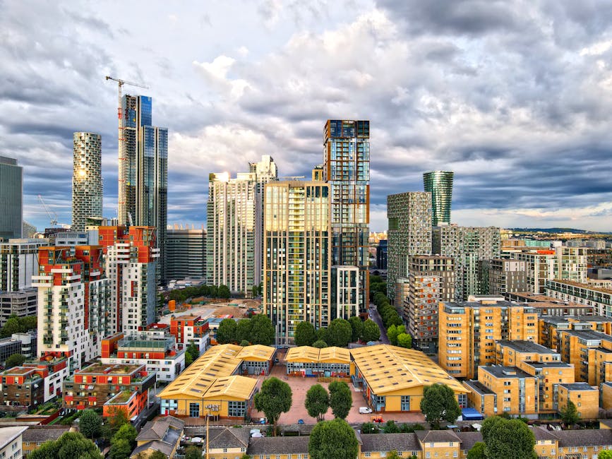 A wide view of a modern city skyline with numerous high-rise buildings, including some under construction with cranes, and a mix of glass and concrete facades. In the foreground, there are mid-rise residential and commercial buildings with flat rooftops, surrounded by green trees and small streets. The scene is captured during daytime with a partly cloudy sky, displaying bright sunlight illuminating the buildings. The image depicts an urban environment suitable for professional house and office removals, with Croydon Man and Van staff possibly involved in moving and transport services related to city relocations or furniture transport within this densely built area.