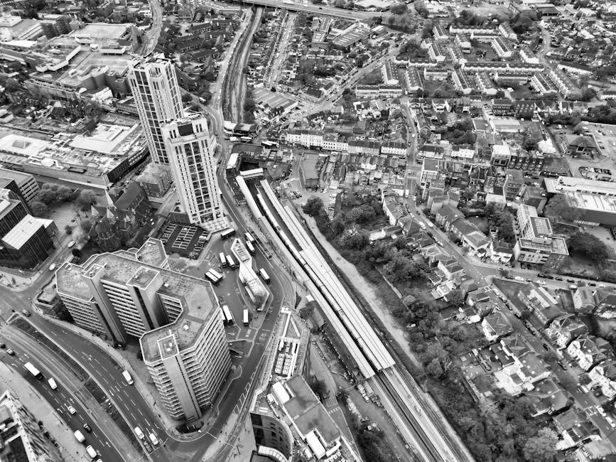 Black and white aerial photograph of a dense urban area toward Croydon, showing a mix of tall office buildings, residential houses, and commercial structures. Prominent office blocks with multiple floors are situated near the foreground, some with flat rooftops, alongside smaller buildings and houses spread out across the landscape. A railway line runs diagonally through the image, with a station platform and several trains visible being loaded or unloaded. Roads with visible traffic, including buses and cars, weave through the cityscape, with one major road in the lower part of the image exhibiting a layered overpass or junction. The scene includes gaps of greenery and trees lining streets and between properties. The environment appears well-lit, with shadows indicating a daytime setting. This aerial view captures the complex transportation infrastructure and residential layout typical of central Croydon, relevant to the logistics involved in house and office removals, as conducted by Croydon Man and Van.