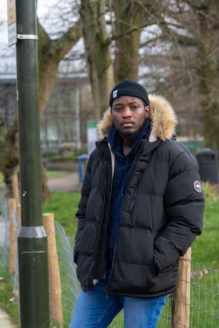 A young man with dark skin, dressed for cold weather, is standing outdoors beside a wooden fence and a metal pole. He is wearing a black padded jacket with a fur-lined hood, a navy blue hoodie underne