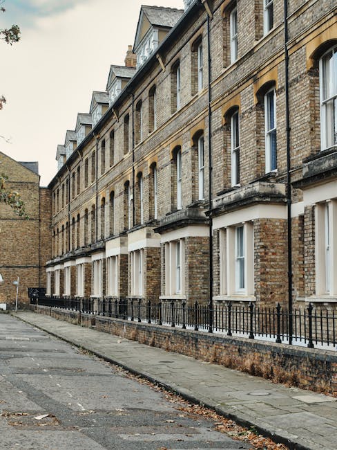 A row of multi-story Victorian terraced houses with brick facades and white-framed sash windows, situated along a narrow pavement in South Croydon. The houses feature decorative stone lintels and small balconies above the ground-floor windows. In front of the properties, a low black metal fence runs parallel to the pavement, separating the houses from the street. The scene is illuminated by natural daylight, with some fallen leaves scattered on the pavement. On the quiet street, a black van from Croydon Man and Van is parked near the curb, with its rear doors open, indicating the beginning of a home relocation process. Nearby, a trolley loaded with unpacked cardboard boxes, wooden furniture parts, and plastic-wrapped items is being prepared for transport or loading. This image captures the logistical elements involved in packing and moving home within the context of house removals and furniture transport services in South Croydon.
