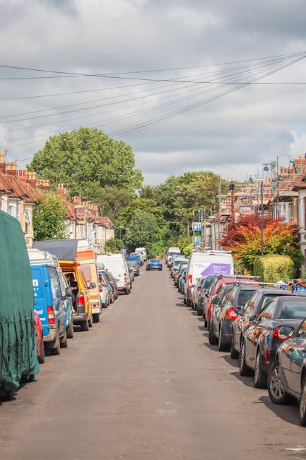 A residential street scene with parked cars lining both sides of the narrow Victorian terrace homes in South Croydon, featuring a variety of vehicles including small vans, larger moving trucks, and private cars, with some of the vans covered in protective fabric or plastic wraps for furniture transport during a home relocation. The pavement is free of debris, and the street extends into the distance, with lush green trees and well-maintained garden plants visible in front of some houses. Overhead, electrical wires crisscross the cloudy sky, and the area is well-lit by natural daylight. This environment reflects a typical setting for a furniture transport and packing and moving process, with the vehicles and arrangements indicative of professional removals services, such as those provided by Croydon Man and Van, specialising in house removals in the area.
