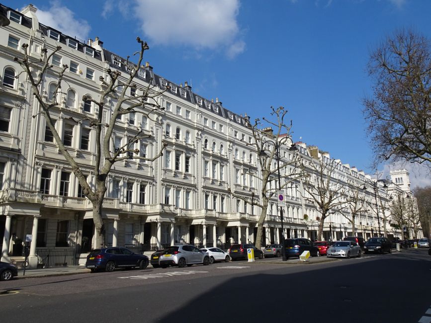 A row of multi-story Victorian terraced houses with brick facades and white-framed sash windows, situated along a narrow pavement in South Croydon. The houses feature decorative stone lintels and small balconies above the ground-floor windows. In front of the properties, a low black metal fence runs parallel to the pavement, separating the houses from the street. The scene is illuminated by natural daylight, with some fallen leaves scattered on the pavement. On the quiet street, a black van from Croydon Man and Van is parked near the curb, with its rear doors open, indicating the beginning of a home relocation process. Nearby, a trolley loaded with unpacked cardboard boxes, wooden furniture parts, and plastic-wrapped items is being prepared for transport or loading. This image captures the logistical elements involved in packing and moving home within the context of house removals and furniture transport services in South Croydon.