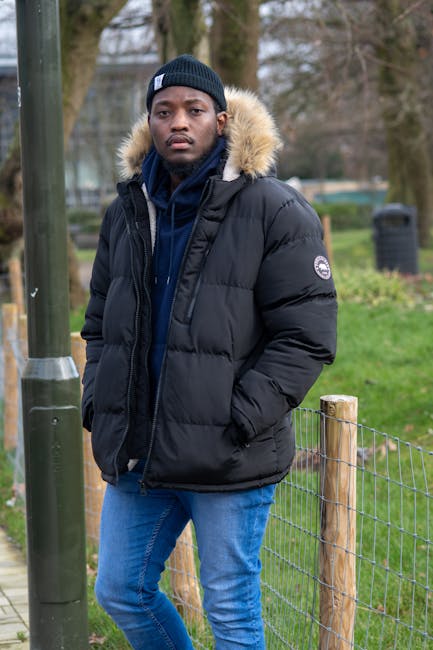 A young man with dark skin and an average build standing outdoors in a park-style setting during daytime. He is wearing a black puffer jacket with a fur-lined hood, a navy blue hooded sweatshirt, and a matching black beanie hat. His hands are in the pockets of his jacket. Behind him, there is a small wooden fence with wire mesh, a metal pole, and trees with bare branches, suggesting a winter or early spring atmosphere. The grass is green, and there are some shrubs and leafless trees in the background. This image could relate to house removals or furniture transportation, illustrating a person preparing for a home relocation process, with the outdoor environment setting the scene for logistical or packing activities involved in moving services provided by Croydon Man and Van.
