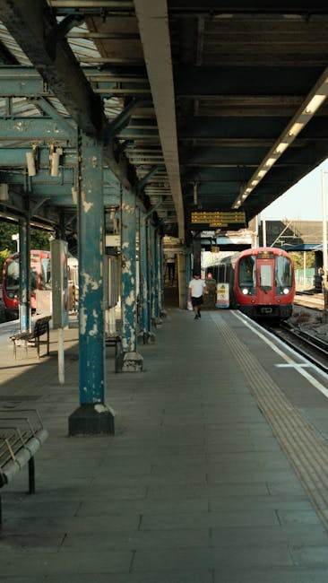 Image of a train platform at Croydon station with a red train arriving or departing on the right side, and a single passenger walking along the platform wearing a light-colored shirt and dark trousers. The platform is sheltered by a weathered metal canopy supported by painted blue steel columns with peeling paint. Benches and small trash bins are positioned along the waiting area, and tactile paving runs parallel to the train tracks for accessibility. The atmosphere appears calm with natural daylight illuminating the scene, emphasizing the station's utilitarian design. This setting reflects the typical environment for home relocation or moving logistics involving train transport, as seen in services by Croydon Man and Van, which support efficient furniture transport, packing, and loading processes.