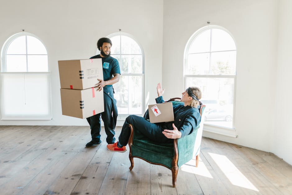 A professional movers from Croydon Man and Van are engaged in a home relocation process inside a well-lit room with white walls and large arched windows providing natural daylight. One mover, standing near the center of the room, is holding two cardboard boxes, one stacked on top of the other, secured with red and black packing tape; he has a friendly expression and is dressed in dark work clothes. The second mover is seated comfortably in a vintage-style armchair with wooden legs and teal upholstery, holding a smaller cardboard box with a red arrow sticker, and is gesturing with one hand as if coordinating or explaining the next step. The wooden floor is clear of debris, and the scene captures the unloading and packing process typical of furniture transport and boxing during a house move. The open space and the presence of boxes suggest an ongoing packing or loading operation associated with a professional removals service.
