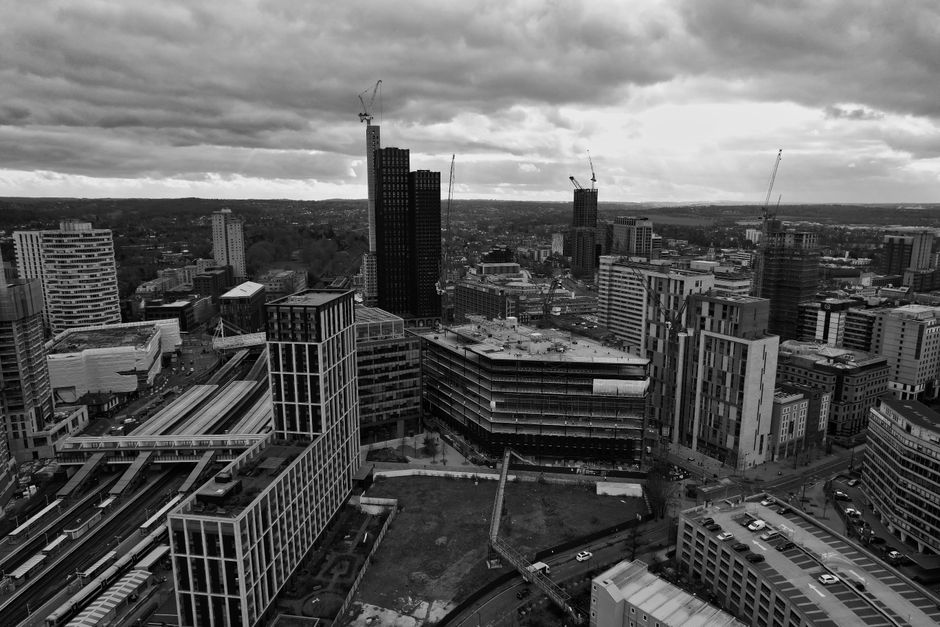A black and white aerial photograph of a cityscape featuring numerous high-rise buildings under construction, with cranes visible atop the tallest structures. The scene includes modern office towers and commercial buildings with glass facades, alongside a network of roads and railway tracks. In the foreground, there is a construction site with some empty space and a crane, possibly indicating ongoing development. The overcast sky with thick clouds casts a diffuse light over the scene. This image exemplifies an urban environment possibly in the process of expansion or renewal, relevant to house removals and relocation services like those offered by Croydon Man and Van for home relocation and furniture transport in the Croydon area.